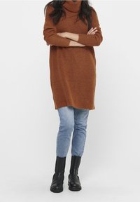 Robe pull en tricot marron avec col roulé et manches longues, associée à un jean ajusté bleu clair et des bottines noires. La texture semble douce.
