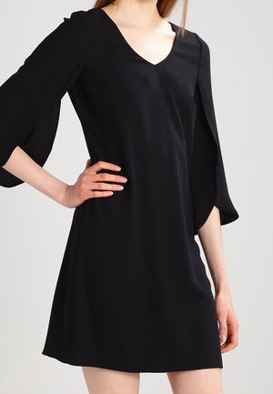 Frau, die ein schwarzes V-Ausschnitt-Kleid mit dreiviertellangen, geteilten Glockenärmeln trägt, steht mit einer Hand in der Hüfte vor einem schlichten Hintergrund.