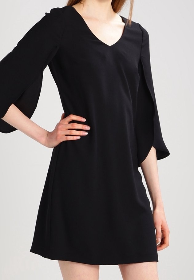 Femme portant une robe noire à encolure en V avec des manches évasées fendues aux trois quarts, debout avec une main sur la hanche devant un fond uni.