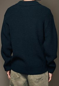Un pull tricoté épais bleu marine avec une texture côtelée, un large col et un design à épaules tombantes. Le dos présente un léger détail de couture.