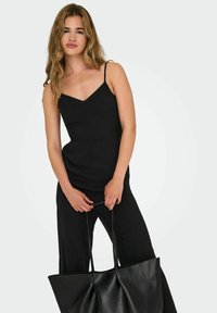 Zwarte jumpsuit met dunne bandjes, V-hals en een comfortabele pasvorm. Model houdt een grote zwarte leren tote bag vast met een zachte textuur en een brede opening.