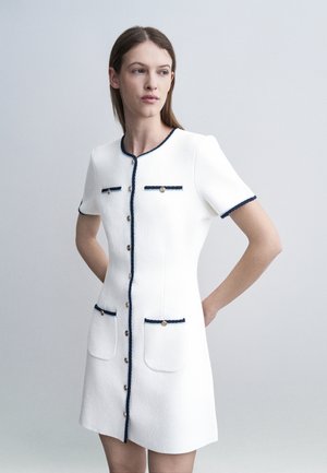 Ženska v beeli obleki s kratkimi rokavi, obrobo v mornarsko modri barvi in gumbi, stoji na enobarvni svetlo sivi podlagi z rokami za hrbtom.