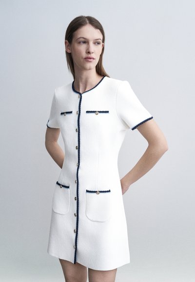 Femme en robe blanche à manches courtes avec bordure et boutons bleu marine, debout contre un fond gris clair uni, les mains derrière le dos.