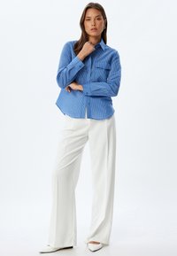 Camisa de botones a rayas azul claro con un bolsillo en el pecho, combinada con pantalones blancos ajustados y zapatos blancos de punta. Textura de tela suave.