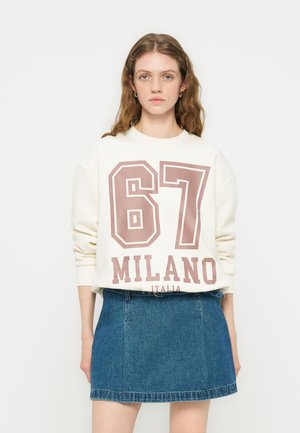 Vrouw draagt een wit sweatshirt met de tekst "67 MILANO ITALIA" in grote letters en een blauwe denim rok, staand tegen een effen achtergrond.