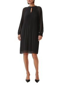 Schwarzes, plissiertes Kleid mit einem Schlüssel-Ausschnitt, langen transparenten Ärmeln, mittellangem Schnitt und strukturiertem Stoff. Getragen mit schwarzen, spitzen Absatzschuhen.