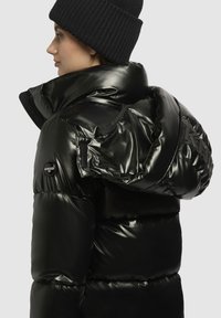 Svart puffjacka med en blank, vadderad ytfinish. Har en hög krage och fast huva, med en logotypdetalj på ärmen.