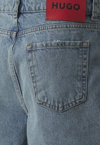 Lyseblå denim jeans med en rød skinnlapp merket "HUGO." Har standardlommer og kontrastsømmer langs sømmene.