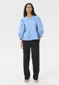 Blusa azzurro chiaro a maniche puff con scollo a V e orlo scallop, abbinata a pantaloni larghi a righe nere e sandali piatti.
