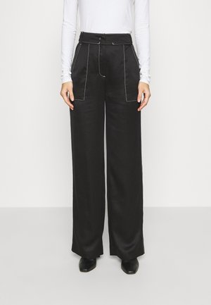 Osoba nosząca czarne, szerokie spodnie z wysokim stanem, z białym przeszyciem i przednimi kieszeniami, w połączeniu z dopasowaną, białą bluzką z długim rękawem oraz czarnym obuwiem.