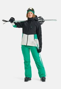Skiausrüstung mit einer schwarz-weißen Farbblockjacke mit grünen Akzenten, kombiniert mit grünen Hosen. Zubehör umfasst Handschuhe und eine Skibrille.