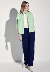 Ljusgrön skjorta med knappar, vit skjorta under, navyblå vida byxor och vita sneakers. Slät tygkvalitet med en avslappnad passform.