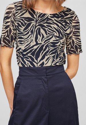 Femme portant un haut beige et noir à motif de feuilles avec des manches courtes et un pantalon taille haute bleu marine foncé sur un fond neutre.