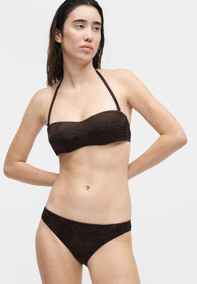 Γυναίκα με μακριά σκούρα μαλλιά, φορώντας ένα καφέ σκούρο μπικίνι με υφή σε δύο κομμάτια, όρθια με τα χέρια πίσω από την πλάτη της, μπροστά σε απλό ανοιχτό φόντο.