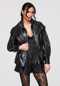 Mujer con chaqueta de cuero negra oversize, top negro, shorts de cuero y medias de encaje floral, posando contra un fondo claro.