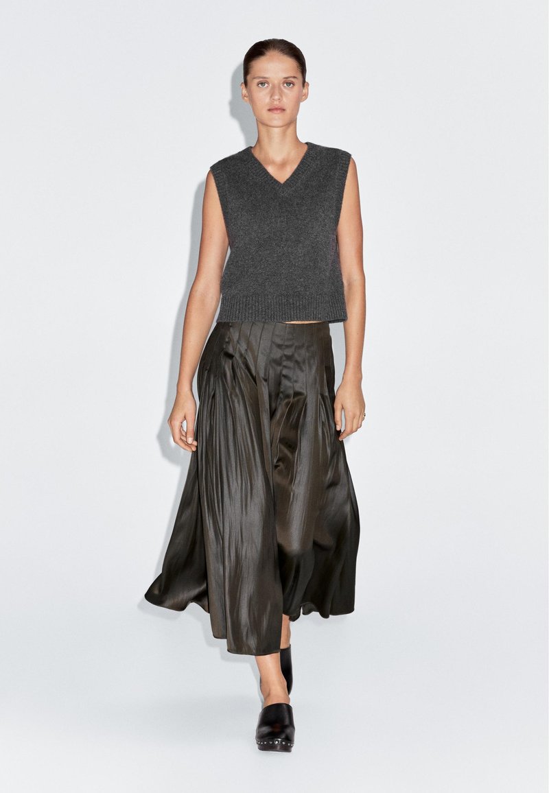 Šedý bezramínkový pletený top s výstřihem do V, spárovaný s plisovanou sukni z tmavě olivového saténu. Model má na sobě černé dřeváky. Jednoduché pozadí.