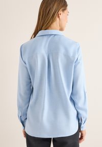 Camicia a maniche lunghe blu chiaro con collo a camicia, dettaglio della piega sul retro e polsini con bottoni, che mette in mostra un tessuto liscio e testurizzato.