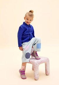 Blaue Jeansjacke, pastellgestreifte Hose mit pinken und blauen Patches und graue Schuhe mit lila Akzenten, die auf einem gepunkteten Hocker sitzen.