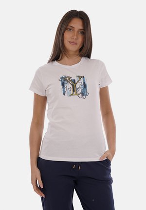 Donna con capelli castani lisci che indossa una maglietta bianca a maniche corte con una grande "Y" e un motivo floreale, abbinata a pantaloni blu scuro.