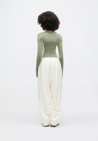 Γυναίκα με σγουρά μαλλιά, φορούν στενό μακρυμάνικο τοπ σε ελιά πράσινο και φαρδύ λευκό παντελόνι, στέκεται με την πλάτη σε απλό φόντο.