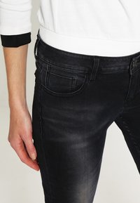 Jeans de mezclilla negros con un diseño ajustado, que cuentan con bolsillos frontales y una textura suave. Una parte de una camiseta blanca con detalles en negro es parcialmente visible.