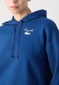 Felpa con cappuccio blu in tessuto morbido. Presenta il nome del marchio ricamato in bianco e un logo alligatore sul lato sinistro del petto.