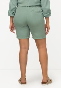Grønne shorts med elastisk talje, glat tekstur og en baglomme. Båret med brune slip-on sko, der har dekorative detaljer.