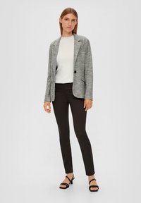 Blazer gris à motifs avec revers, fermeture à un bouton, porté sur un haut blanc, associé à un pantalon noir slim et des sandales à talons noires.