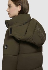 Green puffer jacket with a high collar, quilted texture, and side zipper pockets. Features a circular logo and raised text on the sleeve.
