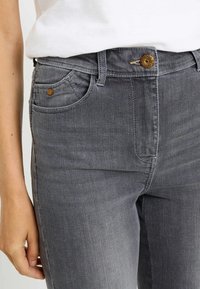 Vaqueros de mezclilla gris con diseño ajustado, estilo de cinco bolsillos y cierre de botón en tono dorado en la cintura. Textura suave.