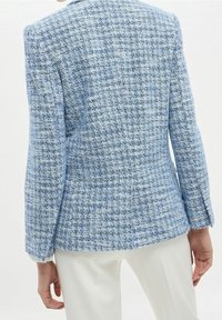 Světlomodrý tweedový kabát s texturovaným vzorem, pouzdrovým střihem a dlouhými rukávy, kombinovaný s bílými kalhotami.