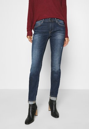 Person wearing cuffed dark blue skinny jeans, black heeled ankle boots, and a long-sleeve red top, standing against a white background.