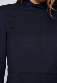 Námořnické šaty s vysokým výstřihem, hladkou texturou, strukturovaným designem a čistými liniemi. Látka vypadá mírně texturovaně se švy detailů.