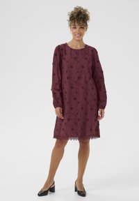 Burgundy klänning med långa ärmar, blommönster, spetsdetalj vid nederkanten, lös passform och rund halsringning, kombinerad med svarta skor.