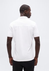 Άνδρας που στέκεται πλάτη φορώντας ένα απλό λευκό πόλο με κοντά μανίκια και μαύρο παντελόνι μπροστά σε λευκό φόντο.