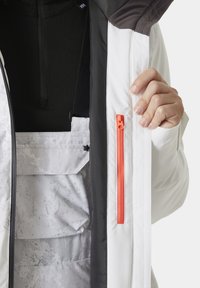 Hvit jakke med svart innside og oransje glidelås, med teksturert fôr og en lomme med klaff. Hender viser detaljene på plagget.