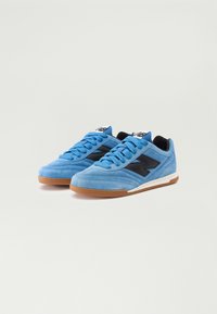 Adidași din suede albastru cu accente negre, bot rotund, talpă plată din cauciuc și șireturi texturate. Prezintă un logo pe limbă și pe lateral.