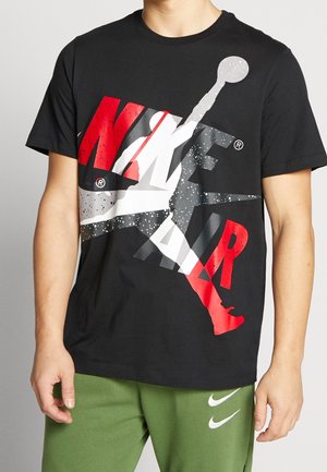 Personne portant un t-shirt Nike Air noir avec un logo Jumpman rouge, blanc et gris et un short Nike vert olive avec des logos Swoosh blancs.