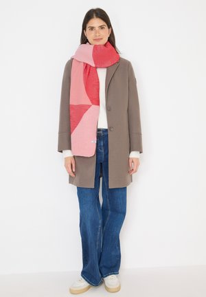 Sciarpa lavorata a maglia con tonalità di rosa e rosso e motivi geometrici, indossata su un maglione crema e un cappotto grigio, abbinata a jeans azzurri chiari.