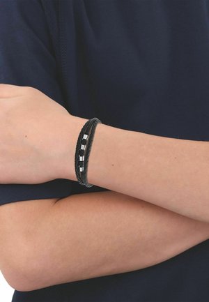 Persona que lleva una pulsera de cuero trenzado negra con cuentas rectangulares de plata en la muñeca, con los brazos cruzados sobre una camisa azul marino oscuro.