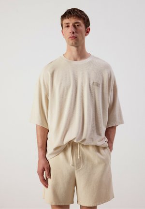 Giovane uomo con camicia beige oversize a maniche corte e pantaloncini abbinati con coulisse, in piedi davanti a uno sfondo chiaro e uniforme.