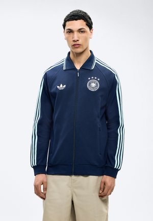 Νεαρός άνδρας που φοράει μπλε σκούρο ζακέτα με φερμουάρ, με ανοιχτόχρωμες ρίγες στα μανίκια και λογότυπα ποδοσφαιρικής ομάδας στο στήθος, στέκεται μπροστά σε λευκό φόντο.