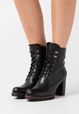 Botines de cuero negro con tacón grueso, diseño con cordones, ojales metálicos y ribete peludo alrededor de la abertura. Suela de goma texturizada.