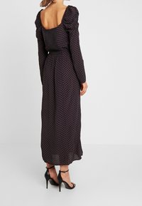 Lång, mörk klänning med puffärmar, små röda prickar, smockad bak och en svävande kjol. Bärs med svarta klackskor med remmar.