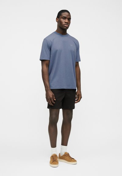 Άνδρας που φοράει μια μπλε σκούρα μπλούζα, μαύρα σορτς, λευκές κάλτσες πλήρους μήκους και καφέ σουέντ αθλητικά παπούτσια, στέκεται μπροστά σε ένα λευκό, απλό φόντο.