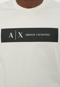 Hvit bomulls t-skjorte med et svart rektangulært grafisk trykk som har "A/X ARMANI EXCHANGE" og logo i hvitt. Korte ermer og rund hals.