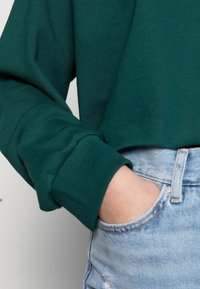 Felpa corta verde scuro con polsini a coste, abbinata a jeans chiari strappati. Il tessuto sembra morbido e dalla consistenza lavorata.