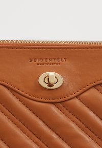 Ruskea nahkainen käsilaukku, jossa on tikattu kuvio, kultasävyinen solki ja vetoketju, edessä yksityiskohtaisesti prässätty logo.