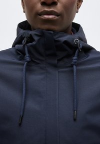 Κοντινό πλάνο ενός ατόμου που φοράει μπλε θαλάσσιο μπουφάν με ψηλό γιακά και ρυθμιζόμενους κορδονέους σφιγμένους στον λαιμό.