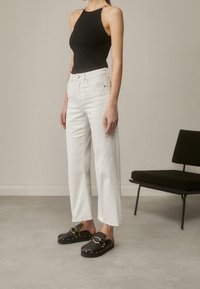 Schwarzes Tanktop, weiße weit geschnittene Jeans mit fransigem Saum und schwarze Clogs mit goldenen Kettenakzenten und Nieten. Minimalistische Sitzgelegenheiten im Hintergrund.